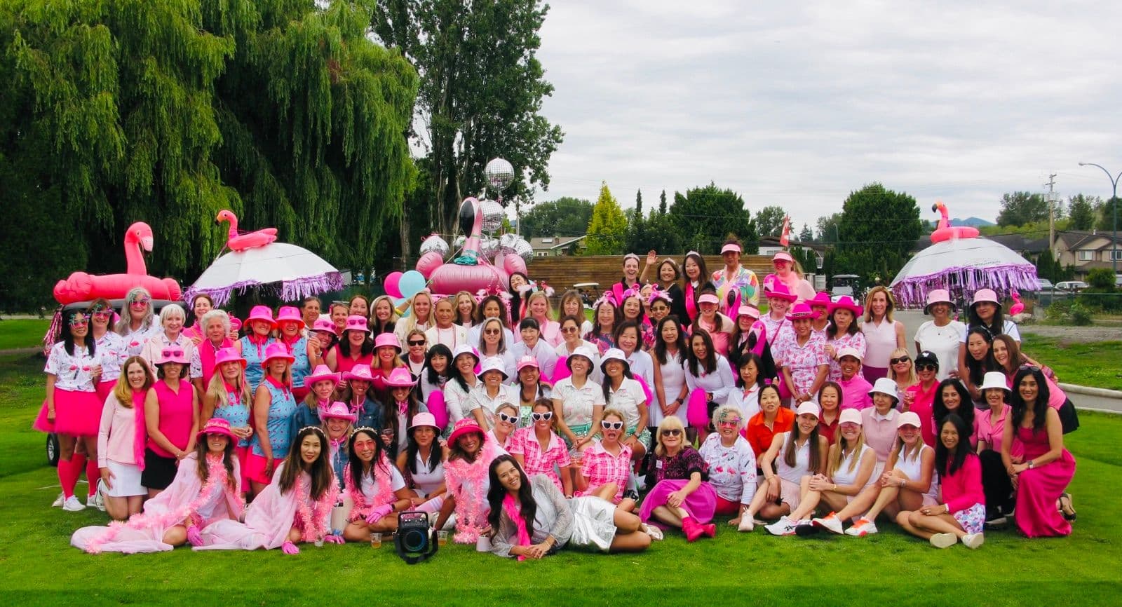 About 90 women dressed in pink pose together on the grass at Musqueam Golf Course, with inflatable flamingos and a decorated golf cart behind them.